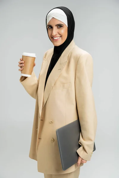 Счастливая и модная мусульманка-бизнесмен с напитком на вынос и ноутбуком, смотрящая в камеру на сером — стоковое фото