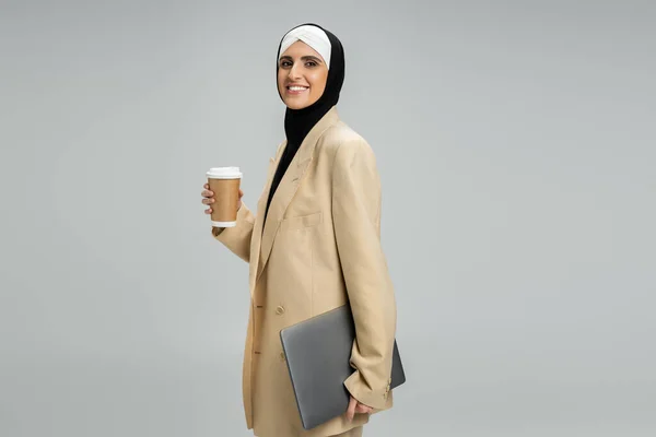 Mulher de negócios muçulmana moderna e elegante com café para ir e laptop sorrindo para a câmera no cinza — Fotografia de Stock