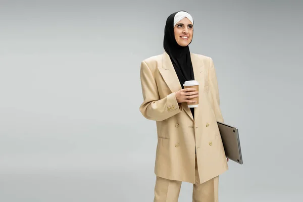 Восторженный и модный мусульманский бизнесмен с бумажной чашкой и ноутбуком, глядя в сторону на серый — стоковое фото
