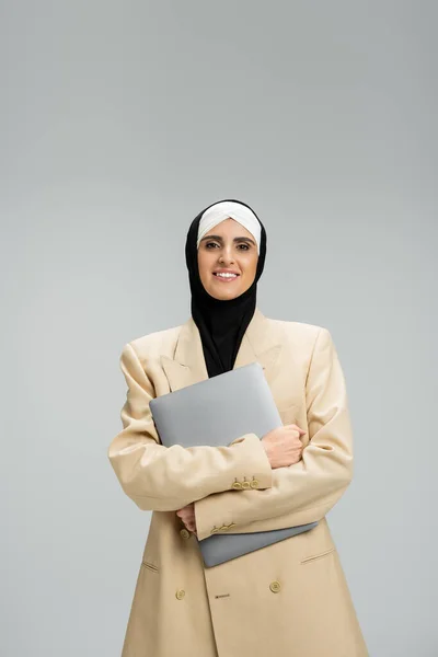 Portrait of smiling muslim businesswoman in blazer and hijab holding laptop on grey, headshot — Stock Photo
