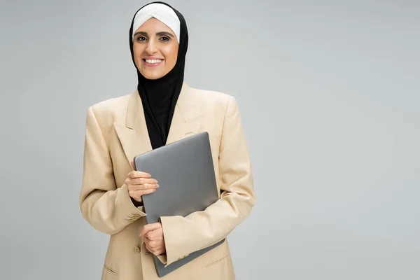 Headshot di donna d'affari musulmana sorridente in giacca e hijab che tiene il computer portatile su grigio, successo — Foto stock