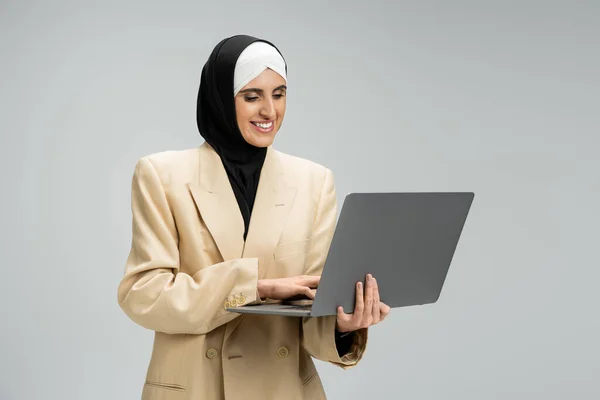 Motivada y alegre musulmana mujer de negocios en hijab y beige blazer networking en el ordenador portátil en gris - foto de stock