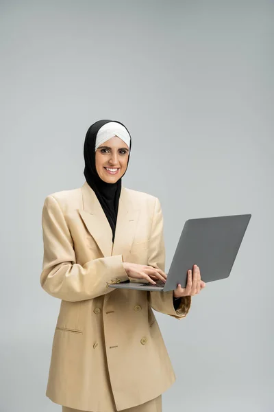 Exitosa mujer de negocios musulmana en hijab y blazer celebración de la computadora portátil y sonriendo a la cámara en gris - foto de stock