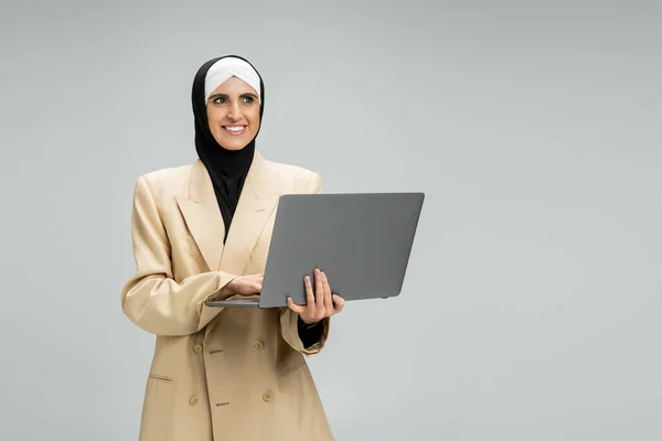 Feliz musulmana mujer de negocios en blazer y hijab celebración de la computadora portátil y mirando hacia otro lado en gris - foto de stock