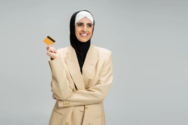 Complacida mujer de Oriente Medio en musulmana hijab y blazer beige con tarjeta de crédito en gris - foto de stock