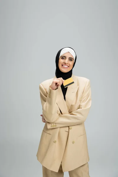 Радостная мусульманка-бизнесмен в хиджабе и костюме держит кредитку и смотрит в камеру на сером — стоковое фото