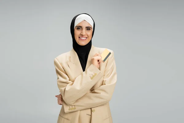 Независимая мусульманская предпринимательница в бежевом пиджаке и хиджабе, стоящая с кредитной картой на сером — стоковое фото