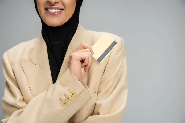 Обрезанный вид улыбающейся мусульманской предпринимательницы в блейзере и хиджабе, держащей кредитную карту на серой — стоковое фото