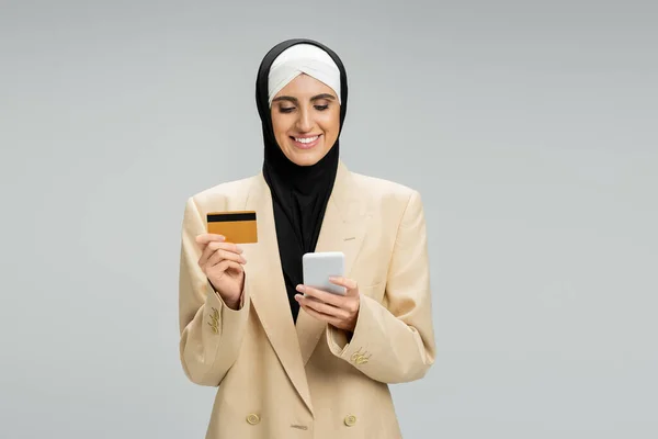 Femme d'affaires musulmane heureux en hijab et blazer élégant tenant smartphone et carte de crédit sur gris — Photo de stock