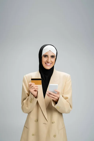 Веселая мусульманка-бизнесмен в хиджабе и модном пиджаке, держащая смартфон и кредитную карту на сером — стоковое фото