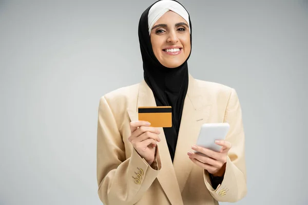 Élégante femme d'affaires musulmane avec téléphone portable et carte de crédit souriant à la caméra sur gris — Photo de stock
