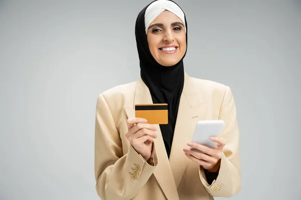 Élégante femme d'affaires musulmane avec téléphone portable et carte de crédit souriant à la caméra sur gris — Photo de stock