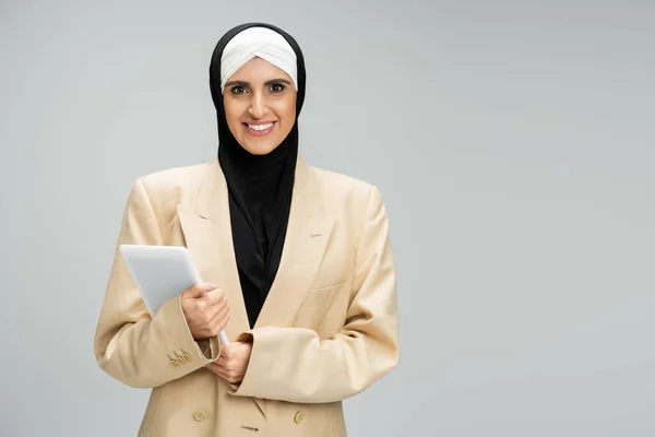 Mujer de negocios musulmana optimista y de moda con tableta digital mirando a la cámara en gris - foto de stock