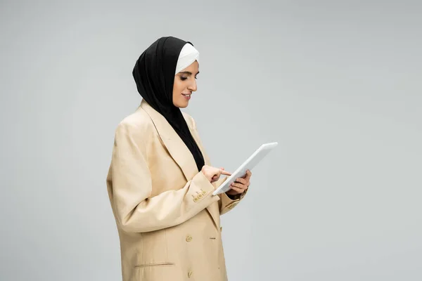 Mujer de negocios musulmana positiva en hijab y beige blazer networking en tableta digital en gris - foto de stock