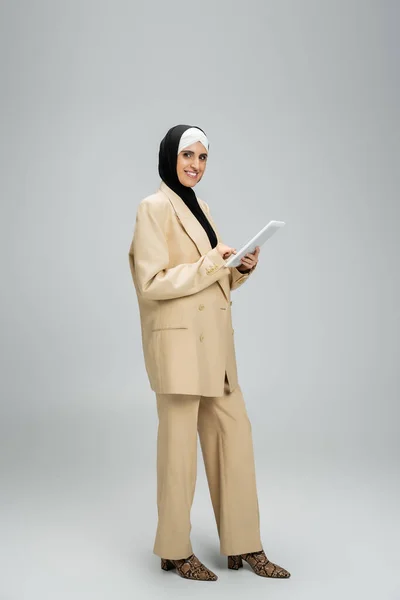 Счастливая мусульманка в бежевом костюме и хиджабе, стоящая с цифровой табличкой на сером, полной длины — стоковое фото