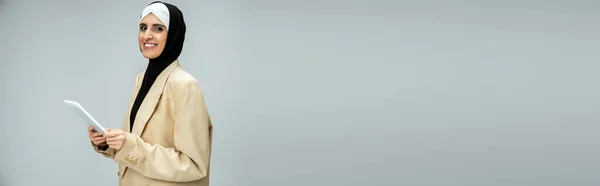 Allegra ed elegante donna d'affari musulmana con tablet digitale in posa su grigio, banner — Foto stock
