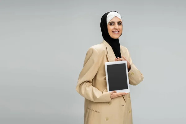 Счастливая мусульманка-бизнесвумен в хиджабе и пиджаке показывает цифровую планшетку с чистым экраном на сером — стоковое фото