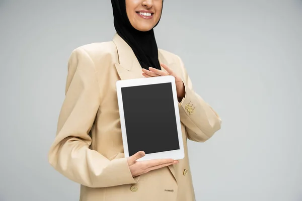 Обрезанный вид улыбающейся мусульманки-предпринимательницы, держащей цифровую табличку с чистым экраном на сером — стоковое фото