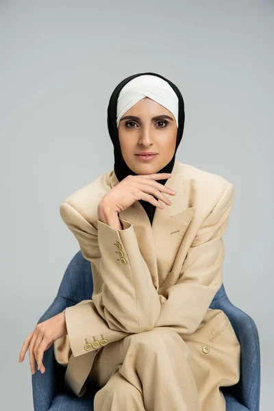 Progressive muslim businesswoman in hijab and stylish suit looking at camera in armchair on grey — Stock Photo