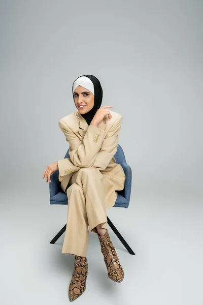 Cheerful muslim businesswoman in stylish suit and hijab smiling at camera in armchair on grey — Stock Photo