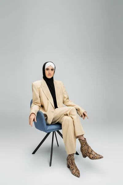 Ambitious muslim businesswoman in trendy suit and hijab sitting in armchair on grey, full length — Stock Photo