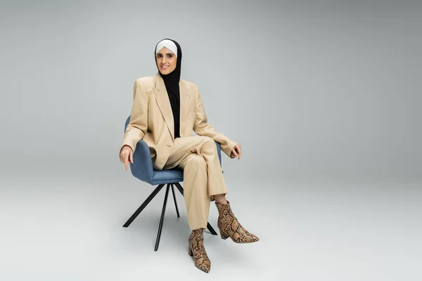 Радостная мусульманка-бизнесвумен в хиджабе и костюме, смотрящая на камеру в кресле на сером, полной длины — стоковое фото