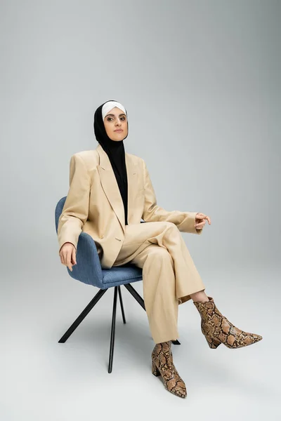 Ambiciosa mujer de negocios musulmana en traje hijab y beige sentada en sillón en gris, bandera - foto de stock