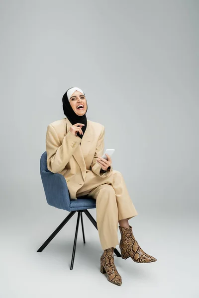 Excited and stylish muslim businesswoman with smartphone sitting in armchair and laughing on grey — Stock Photo