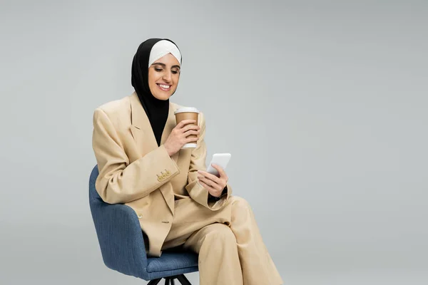 Feliz y de moda musulmana mujer de negocios con teléfono inteligente y café para ir sentado en sillón en gris - foto de stock