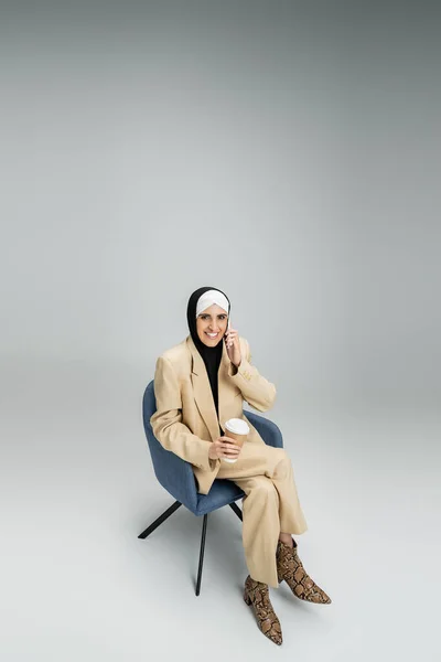 Cheerful, trendy muslim businesswoman with coffee to go talking on smartphone in armchair on grey — Stock Photo