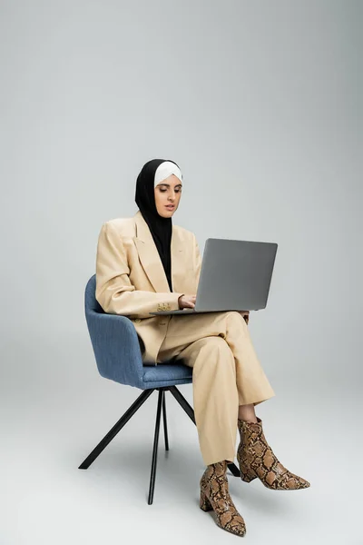 Moderna mulher de negócios muçulmana em hijab e terno trabalhando no laptop em poltrona em cinza, comprimento total — Fotografia de Stock
