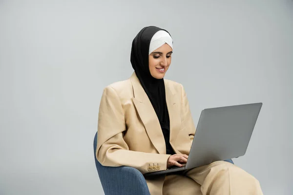Complacida, moderna mujer de negocios musulmana en traje hijab y beige trabajando en el ordenador portátil en sillón en gris - foto de stock