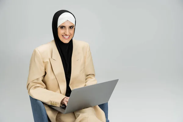 Успешная и модная мусульманка-бизнесмен с ноутбуком, улыбающаяся в камеру в кресле на сером — стоковое фото