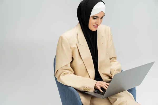 Концентрированная мусульманка-бизнесвумен в костюме и хиджабе, печатающая на ноутбуке в кресле на сером — стоковое фото