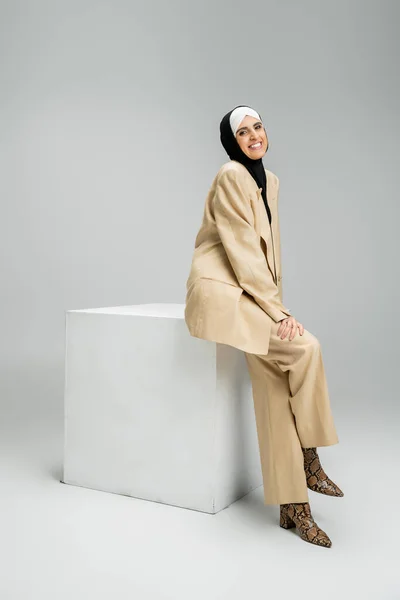Joyful muslim businesswoman in hijab and beige suit sitting on white cube on grey, full length — Stock Photo