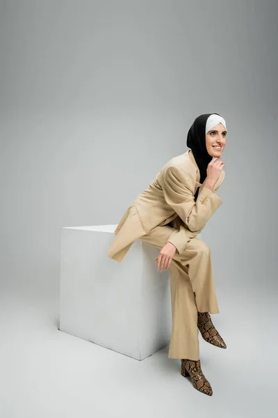 Smiling muslim businesswoman in hijab and elegant suit looking away on cube on grey, full length — Stock Photo