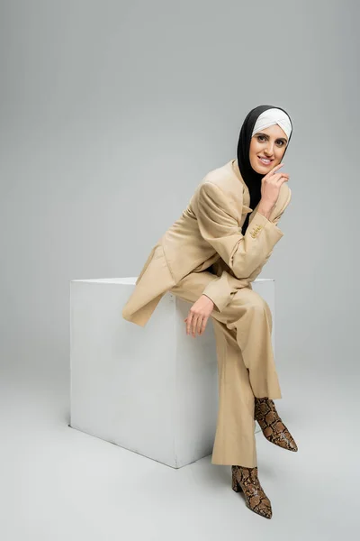 Mujer de negocios musulmana complacida en hijab y elegante atuendo sonriendo a la cámara en cubo blanco en gris - foto de stock