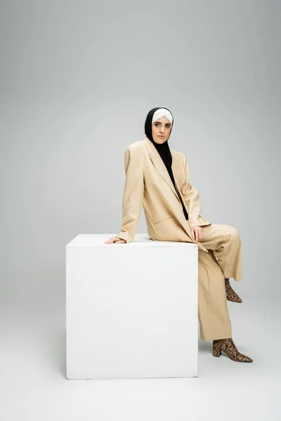 Confident muslim businesswoman in beige stylish suit and hijab sitting on white cube on grey — Stock Photo