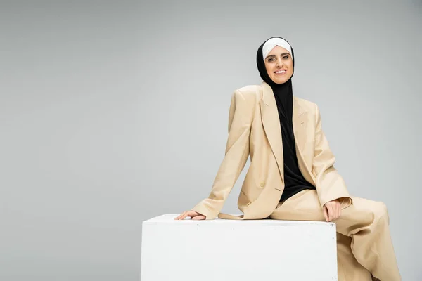 Overjoyed muslim businesswoman in beige trendy suit and hijab sitting on white cube on grey — Stock Photo