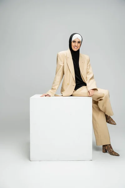 Cheerful muslim businesswoman in fashionable attire and hijab sitting on white cube on grey — Stock Photo