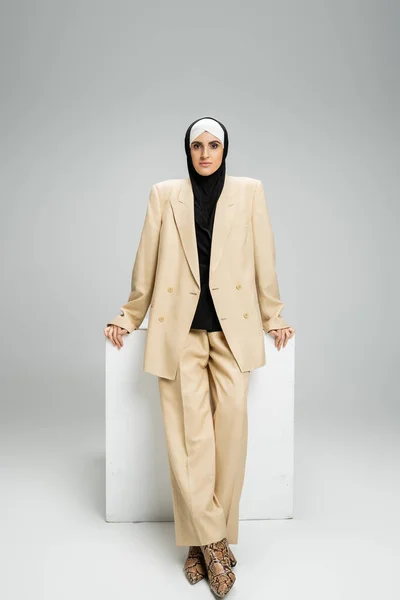 Independent muslim businesswoman in stylish suit and hijab standing near cube on grey, full length — Stock Photo