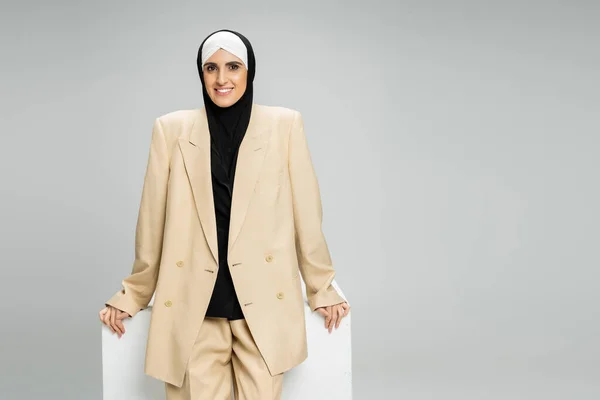 Happy muslim businesswoman in beige suit and hijab smiling at camera near white cube on grey — Stock Photo