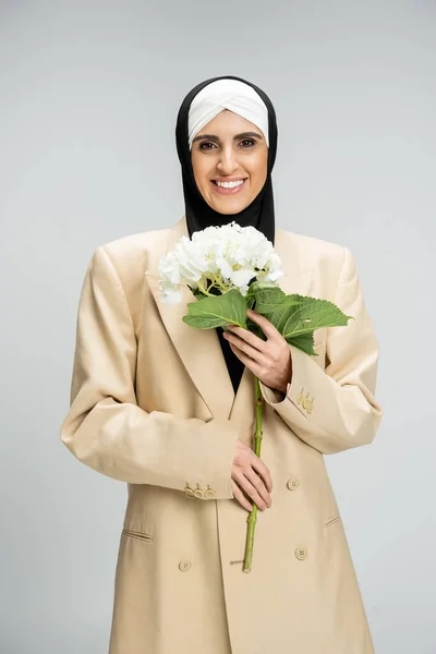 Joyful muslim businesswoman in  hijab and beige blazer holding white hydrangea flower on grey — Stock Photo