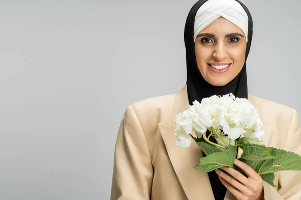 Stylish and cheerful muslim businesswoman with white hydrangea flower and looking at camera on grey — Stock Photo