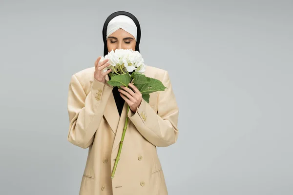 Fashionable muslim businesswoman in hijab and blazer enjoying flavor of hydrangea flower on grey — Stock Photo