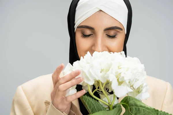 Стильная и современная мусульманская предпринимательница с закрытыми глазами, пахнущая цветком гортензии на сером — стоковое фото