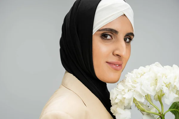 Retrato de mulher de negócios muçulmana elegante com flor de hortênsia branca olhando para a câmera no cinza — Fotografia de Stock
