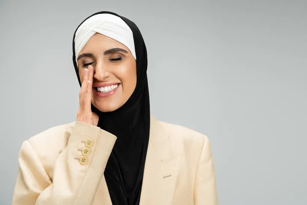 Muslim businesswoman in stylish outfit holding hand near face and smiling with closed eyes on grey — Stock Photo