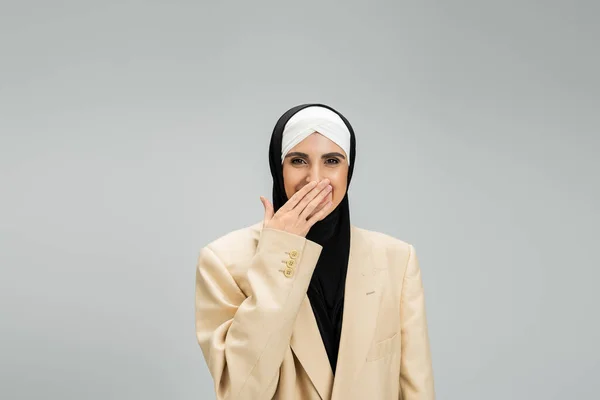 Excited muslim businesswoman in hijab and blazer covering mouth with hand and laughing on grey — Stock Photo
