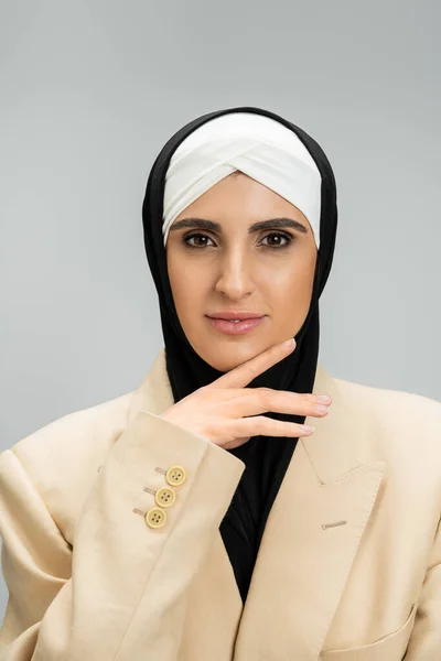 Mulher de negócios muçulmana elegante com maquiagem, em hijab e blazer, de mão dada perto do queixo no cinza — Fotografia de Stock